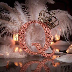 Rose-gold crystal "Q" surrounded by glowing feather wings, a tiny crown sitting atop for a regal finish